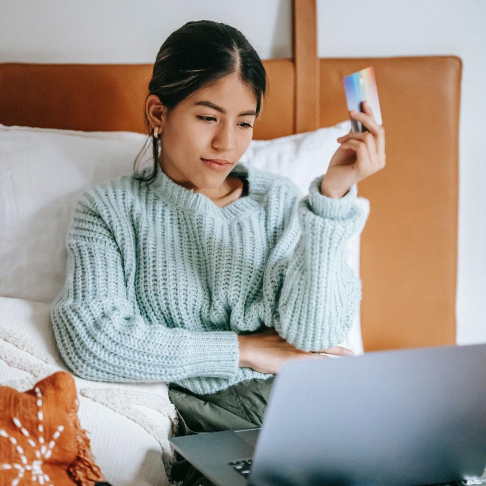 A teen girl in chunky sweater sits in bed and internet shops on her laptop with her credit card help up in one hand, financial literacy