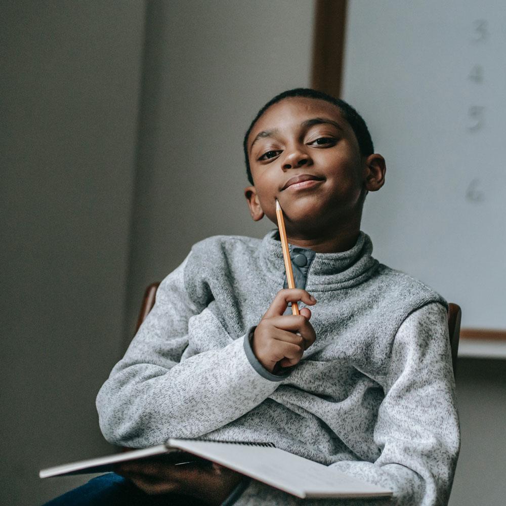 Middle school boy sits in a chair with a classroom markerboard behind him and a notebook and pencil in his hands