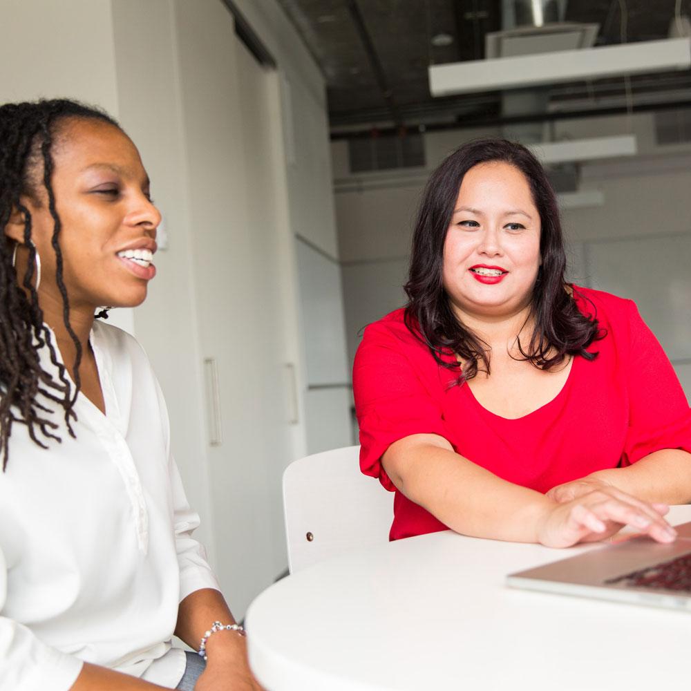 Two diverse women sit at a table and talk about what's on a computer in front of them, teach education